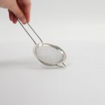 Kleine metalen zeef of sifter voor koken of bakken, handvast gehouden tegen een witte achtergrond.