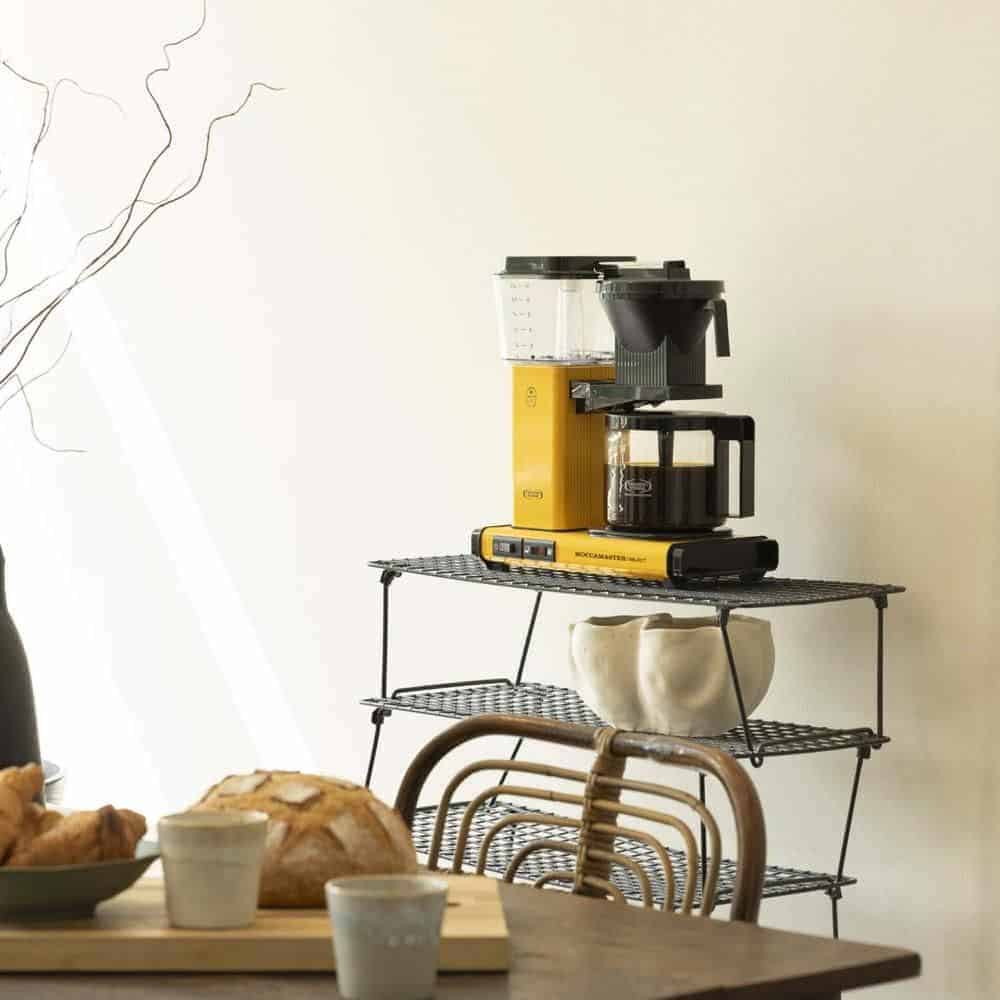 Koffiezetapparaat en accessoires op een metalen rek in een keuken, met brood en kopjes op de tafel eronder.
